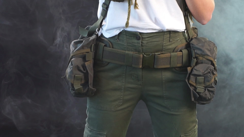 Woman in protective armour and headphones holds gun in hands, posing over dark smoky background. Army, dedication, weapon, technology.