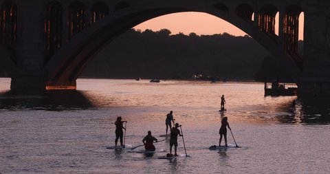 Washington, D.C. tourists explore the Potomac River using stand up paddle boards and kayaks on a hot summer day at sunset. The famous key bridge, a landmark, is in the background.