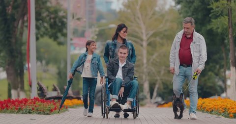 Disabled guy walks with the family in the park. Young man in a wheelchair rides along a path in a public garden.