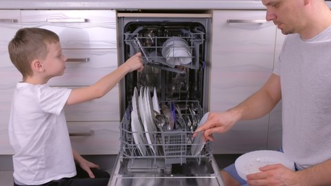 Dad and son are putting a dishes in dishwasher together in the kitchen, Side view.