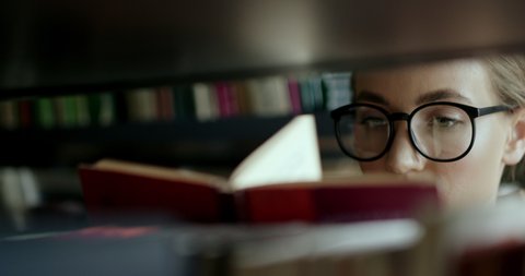Smart woman in glasses taking books from shelf and flipping through pages in library