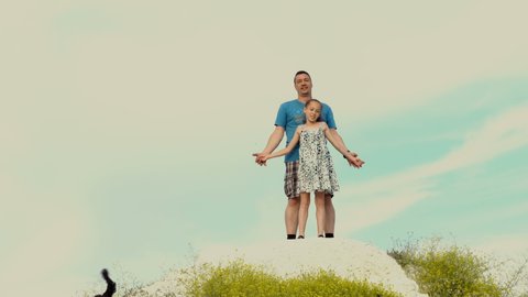 Happy dad and his little daughter are standing on a hilltop on a Sunny day against a bright sky. They hold hands and begin to descend. The concept of a happy family. 4K.