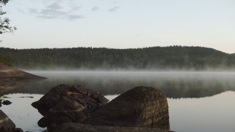 4K anamorphic video of fog mist drifting over beautiful and calm lake fjord with rocks in foreground during sunrise golden hour in Maridalen in Oslo, Norway.