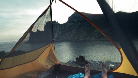 A young guy lies in a camp tent, his legs protrude from the tent. Enjoys relaxing on the beach after the adventure of the trekking day. Camp tent stands on the seashore. Hiking trip in Crimea. Video de stock