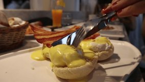 close up video of a fork and knife cutting an egg benedict. Roasted bacon and a fresh fruit juice glass in the background