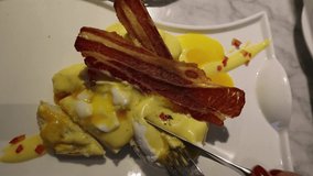 Bird's eye clip of a fork and knife cutting an egg benedict with some roasted bacon