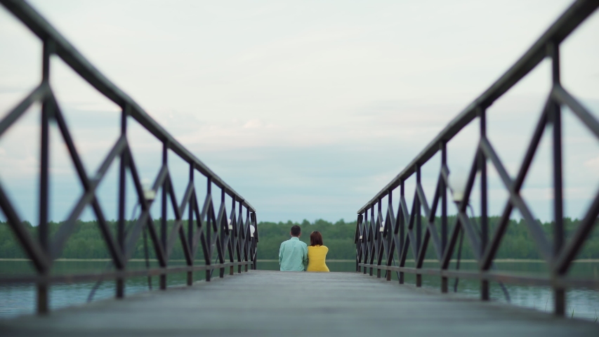 Back perspective view of young loving couple sitting on wooden pier, looking at beautiful river view, talking and embracing warmly