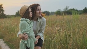 Loving moms puts on daughter knitted blanket, hugging child, summer nature background, golden hour