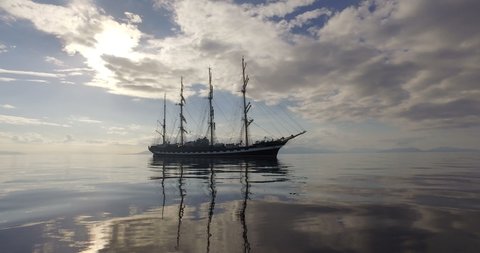 Big Sailing Ship In The Calm Mirror-Smooth Sea On The Background Of The Mountain Coast. The Four-mast Ship Drifts With Lowered Sails On the Sunset.