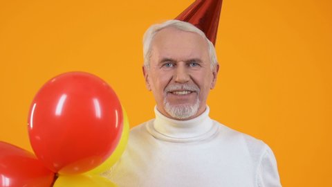 Joyful senior man party hat blowing noisemaker holding balloons, celebration
