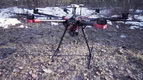 Close up for drone copter standing motionless on the ground in the early spring, video production concept. Clip. Black quadcopter on wet soil with snow and bushes on the background.