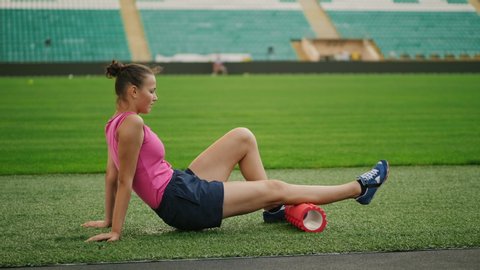 Theme sport and rehabilitation sports medicine. Teen girl athlete uses a massage foam roller sitting on a lawn of a football field in a stadium. Workout to remove pain, stretch and massage muscles