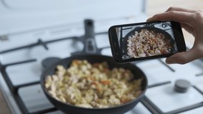 A culinary blog. Woman`s hand with a telephone makes a video of stew vegetables with meat in the fry pan.