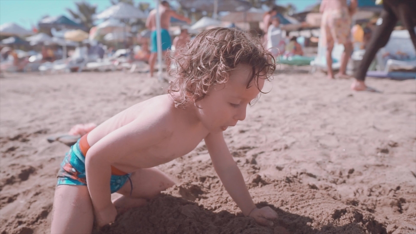 2 years old long haired boy is playing with sand on beach, summertime. Funny children making castle from sand, childhood and summer concept. 
