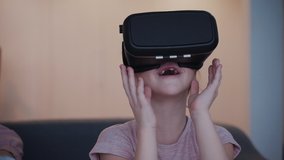 Small girl spending free time playing at games and sitting on comfort sofa in cozy light flat with modern interior room. She watching entertainment program in virtual reality helmet inside her house