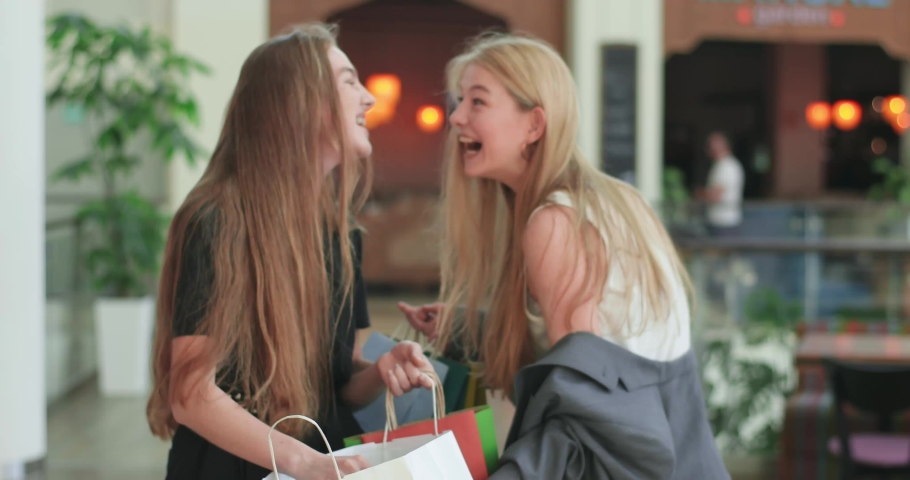 Stylish friends are walking in the mall, laugh and hugging. Happy friends go home after succesful shopping. Two young beautiful woman stroll inside the shopping centre and view their purchases. 4K