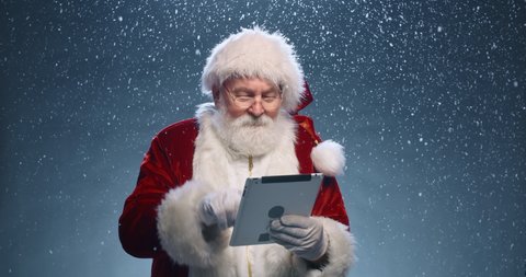 Santa Claus surfing internet on his tablet, then looking at camera nad winking, isolated over snowy blue background - christmas spirit concept close up 4k footage