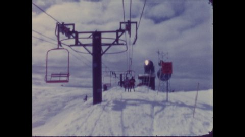 1960s: People ride ski lift to top of mountain, exit ski lift. Mountain, trees, sky, clouds.