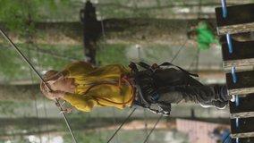 Vertical video. Little boy in a safety harness climbs on a route in treetops in a forest adventure park. He climbs on high rope trail. Outdoor amusement center with climbing activities consisting of