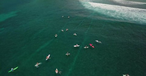 Aerial view of many sporty surfers in clear blue ocean. Camera elevates and follow lucky surfer who can catch good wave on short board. People soaking up the sun waiting for the next big wave.