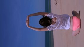 Woman practices yoga sits in lotus pose on sea sandy beach in windy day, back view. She stretches hands up wearing in swimsuit, sunglasses and blouse. Travel tourism on vacation. Vertical video.