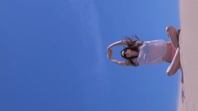 Woman practices yoga sits in lotus pose on sandy beach in windy day. She stretches raises hands up on blue sky background. Swimsuit, sunglasses and blouse. Travel tourism on vacation. Vertical video.