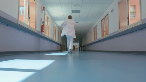 Beginning of the work day. Doctor on the move puts on a white uniform in a hurry and walks down the hospital corridor. Medical worker goes along the corridor of a large modern hospital.