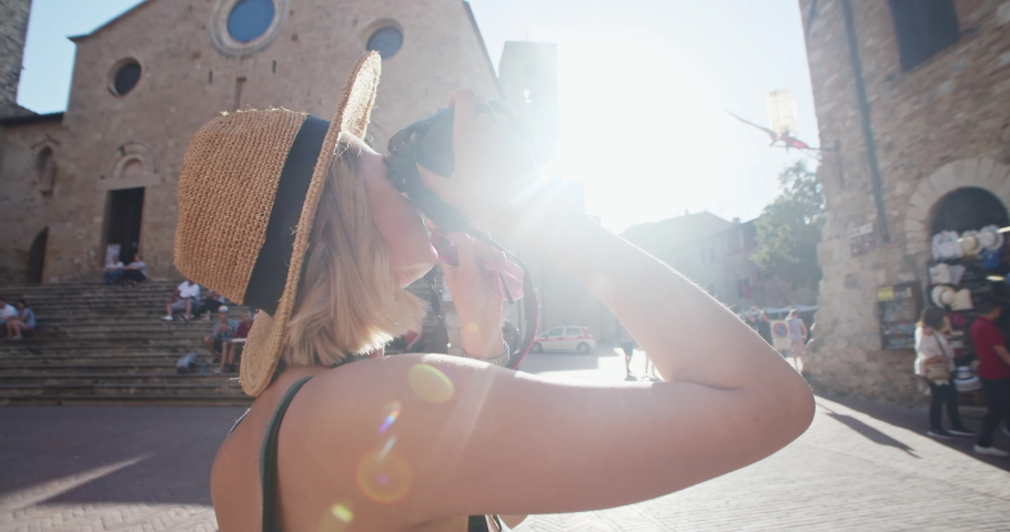 Portrait of amazed good mood blonde girl taking photographs of beautiful historic buildings. Young woman photographer wanderer exploring city during summer journey. Sunrise. Piazza Duomo San Gimignano