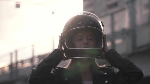 Close-up of young motorcyclist woman taking off retro helmet in the outdoors at sunset. Biker girl in leather jacket dressing for trip. Front view, slow mo