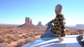 Young man with rental car enjoying road trip in USA ; one man outstretching arms on vehicle hood looking at Monument Valley 