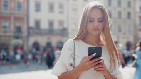 Close up view of young blonde woman in fashionable outfit standing in the city center and actively texting, messaging via her phone, reacts disappointedly and sad. Negative emotions, technology