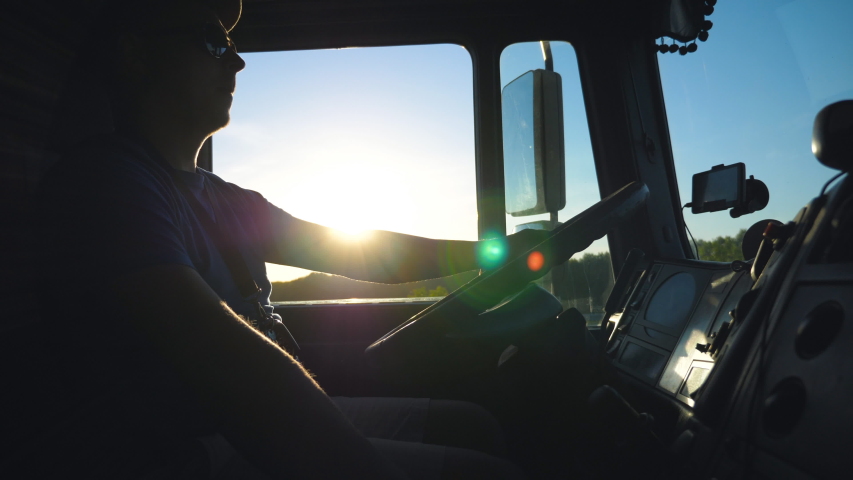 Silhouette of lorry driver driving through countryside in evening. Man in sunglasses controlling his truck attentive watching road. View inside cabin of lorry. Inside shot Slow motion Close up Royalty-Free Stock Footage #1033713842