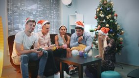 Cheerful friends in Santa hats making video call by smartphone on Christmas party while blowing party whistle, making cheers and having fun together.