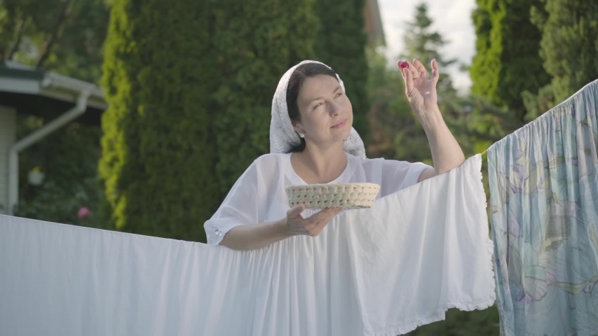 Pretty woman with white shawl on her head eating cherries looking at camera smiling over the clothesline outdoors. Washday. Positive housewife doing laundry. Slow motion.
