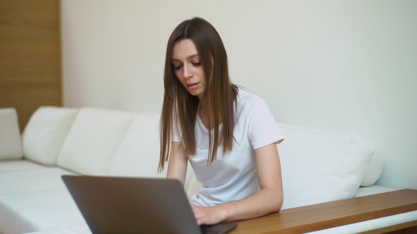 Girl typing on laptop. Guy brings cup of coffee. Young woman drinks coffee. Cute man and female typing on keyboard together sitting on sofa in modern white living room