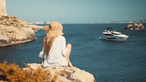 Beautiful happy asian girl in a shirt and wheat hat sitting on the edge of a cliff looking in the sea with a luxury yacht in the background while camera has cinematic movement.
