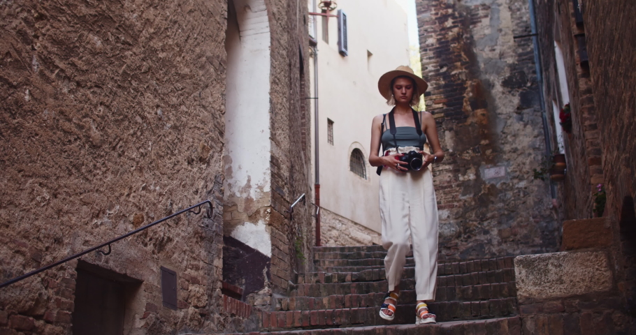 Echanting young woman walking down stairs in wonderful narrow street. Beautiful girl photographer in hat exploring old town of San Gimignano during summer journey. Tuscany, Italy