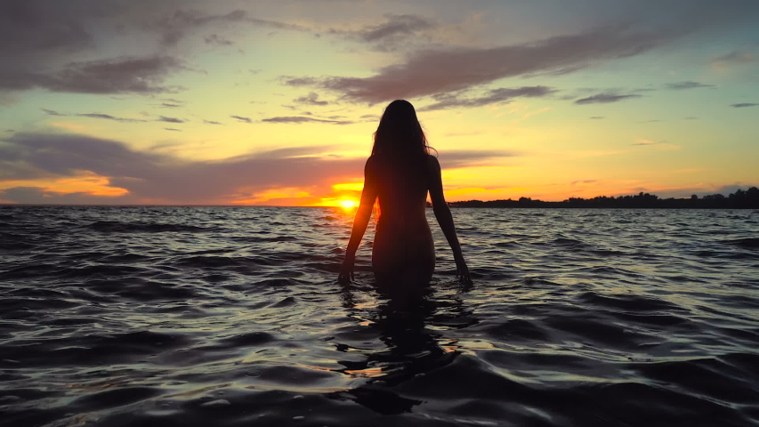 Slim young woman walking in the sea above the sunset sky. Slowmotion. Handheld shot.