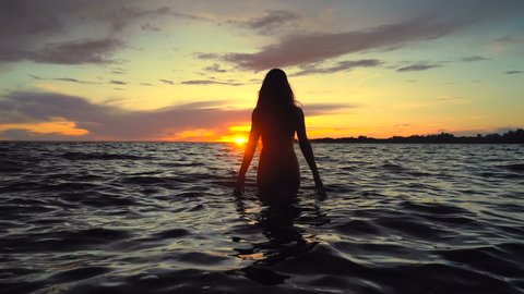 Slim young woman walking in the sea above the sunset sky. Slowmotion. Handheld shot.