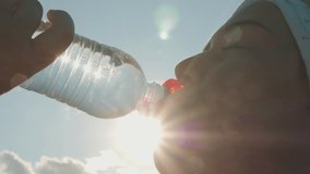 Close-up shot: Overweight girl drinks water from a bottle after a workout outdoors against sun rays and blue sky, slow-motion video