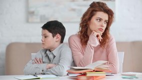 offended preteen boy sitting with crossed arms and looking at mother at desk with books