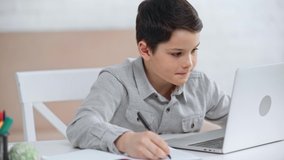 focused preteen schoolboy looking at laptop screen, writing in copy book and typing on keyboard