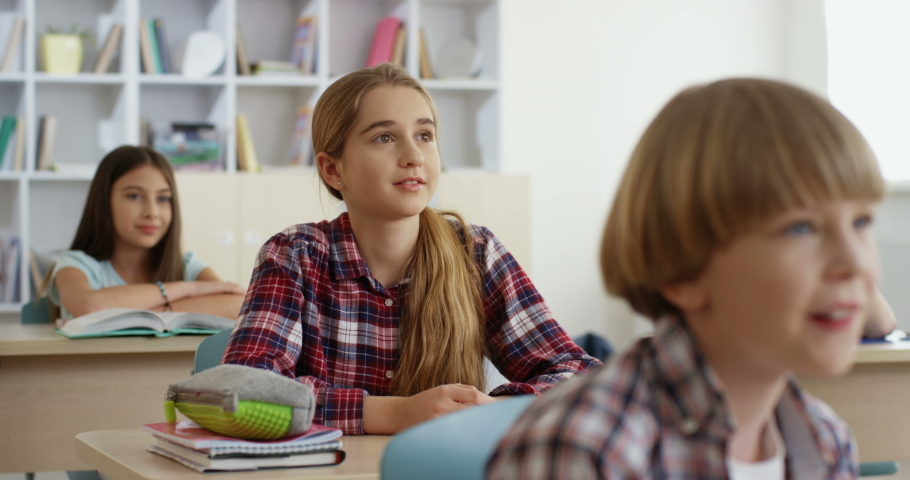 Cheerful blond Caucasian beautiful girl sitting in the classroom at the desk and rising hand as want to answer the question of the teacher during a lesson.