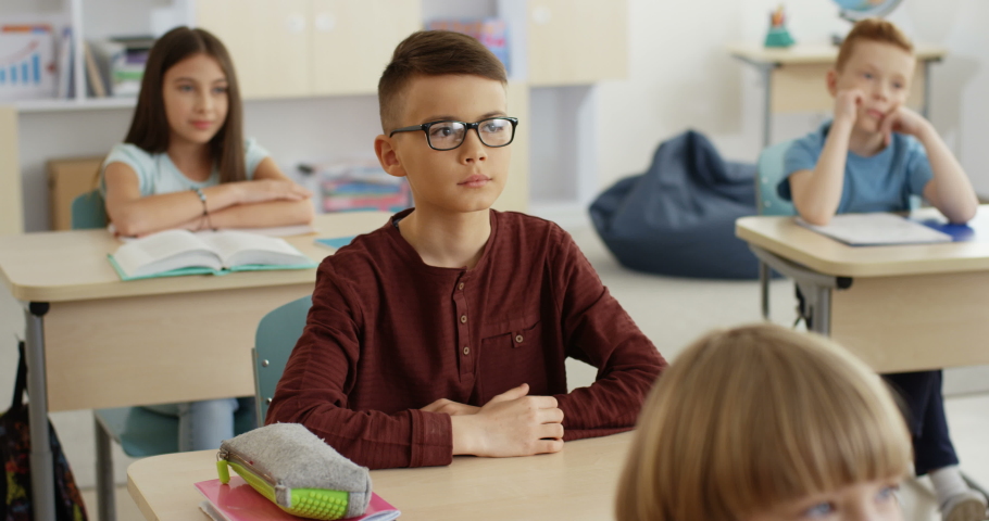 Cute Caucasian small schoolboy in glasses sitting in the classroom at the desk and rising hand as want to answer the question of the teacher during a lesson.