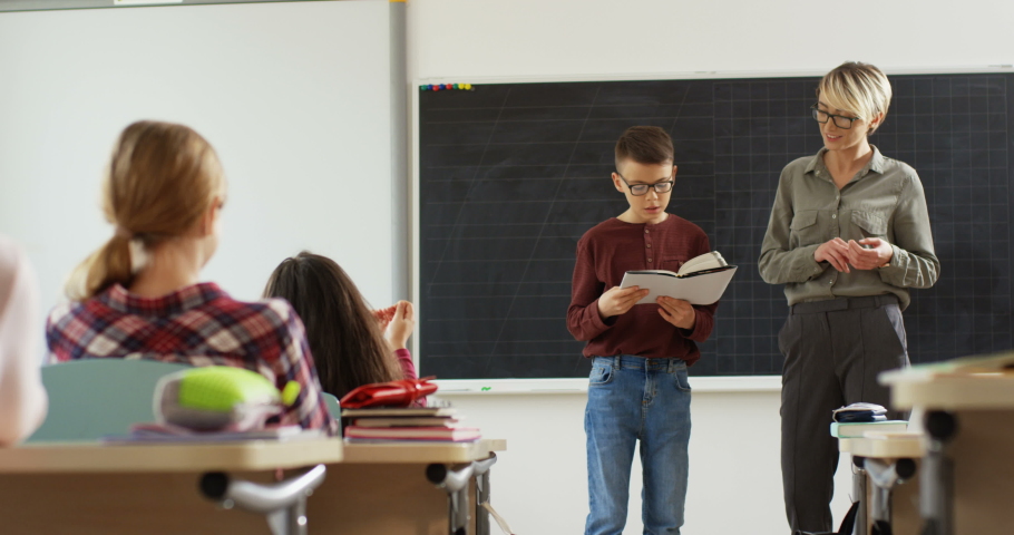 Small teen schoolboy in glasses standing in the middle of the classroom and answering while reading something from the textbook in front of his class and pretty teacher.