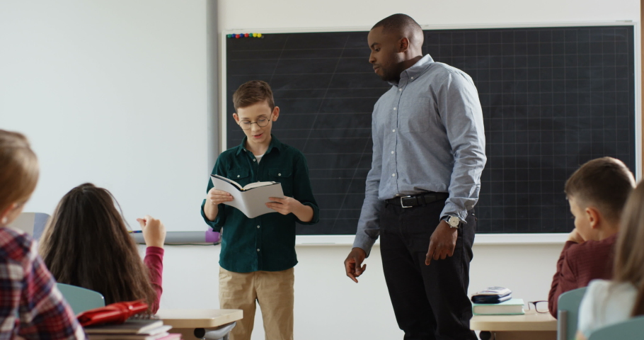 Nice teen schoolboy in glasses standing at the blackboard in front f the class and reading from a textbook to the African American male teacher during literature lesson.