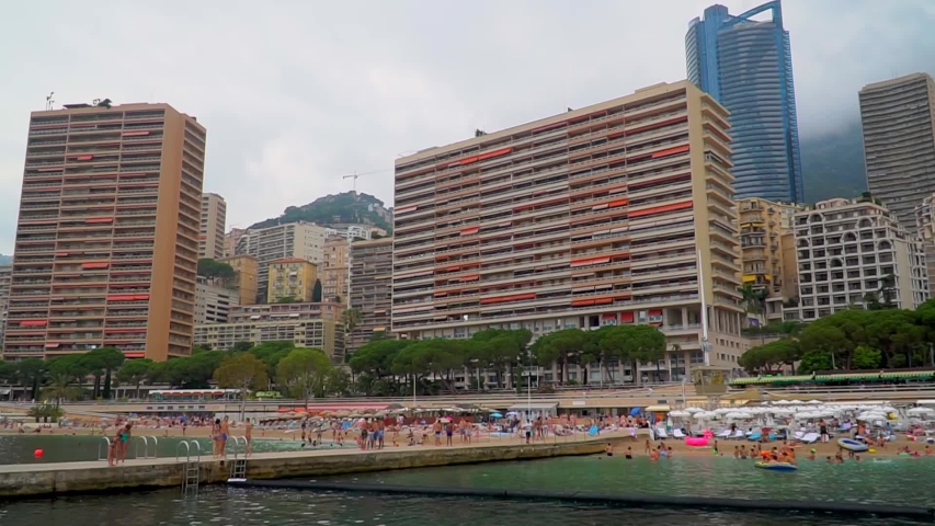 Panorama of the sandy beach of Monaco. Larvotto beach is filled with resting people on sunbeds and in the water against the backdrop of modern architecture of high-rise buildings. Luxury beach
