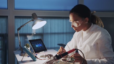 Close up of female engineer of electronics in white coat and protective glasses checking motherboard with multimeter tester and other electronic devises