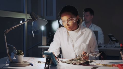 African american electronics engineers checking electronic board with multimeter tester and other electronic devises in lab. There is a fellow worker in the background.