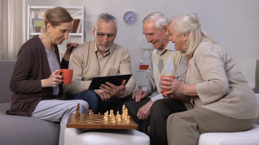Elderly man showing friends new project on tablet pc, chess play brake, company